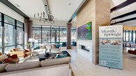 Located at 5401 Atlantic Springs Rd in Raleigh, NC, this single-story clubhouse serves as the social and amenity hub of the Atlantic Springs community. The space is designed with a contemporary aesthetic, showcasing light large-format tile flooring, crisp white walls, and bold exposed black ceiling beams that create a striking visual contrast throughout.

Guests enter through floor-to-ceiling black-framed glass doors that flood the entryway with natural light and offer immediate views of the surrounding community. The main lounge area is anchored by a dramatic wood-clad fireplace column with a large mounted flat-screen display above, flanked by generous seating arrangements and a statement chandelier. Expansive grid-pane windows line the rear wall, providing unobstructed views of the outdoor pool area and bringing the outside in.

Adjacent to the lounge is a stylish café-style bar with white countertops, bar seating, pendant lighting, and a slatted wood accent wall, ideal for casual gatherings. A dedicated coworking alcove features built-in desk space, wall-mounted screens, and large windows, offering a functional workspace with outdoor views. Additional seating nooks with upholstered banquettes provide more intimate settings. The shuffleboard table and open social areas round out a clubhouse designed to foster both relaxation and community engagement.