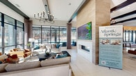 Located at 5401 Atlantic Springs Rd in Raleigh, NC, this single-story clubhouse serves as the social and amenity hub of the Atlantic Springs community. The space is designed with a contemporary aesthetic, showcasing light large-format tile flooring, crisp white walls, and bold exposed black ceiling beams that create a striking visual contrast throughout.

Guests enter through floor-to-ceiling black-framed glass doors that flood the entryway with natural light and offer immediate views of the surrounding community. The main lounge area is anchored by a dramatic wood-clad fireplace column with a large mounted flat-screen display above, flanked by generous seating arrangements and a statement chandelier. Expansive grid-pane windows line the rear wall, providing unobstructed views of the outdoor pool area and bringing the outside in.

Adjacent to the lounge is a stylish café-style bar with white countertops, bar seating, pendant lighting, and a slatted wood accent wall, ideal for casual gatherings. A dedicated coworking alcove features built-in desk space, wall-mounted screens, and large windows, offering a functional workspace with outdoor views. Additional seating nooks with upholstered banquettes provide more intimate settings. The shuffleboard table and open social areas round out a clubhouse designed to foster both relaxation and community engagement.