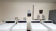 This single-floor shared laundry room is a practical, functional facility designed to serve multiple users. The room is outfitted with commercial-grade Speed Queen washing machines and dryers, arranged in a neat row along the wall for easy access. The machines feature digital control panels, indicating modern, reliable performance suited for high-frequency use. Exposed plumbing pipes run along the wall, providing direct connections to the washers, while dryer exhaust venting is also visible, reflecting a straightforward, utilitarian layout. The walls are finished in a clean, neutral tone, keeping the space bright and easy to maintain. Posted signage on the wall clearly outlines laundry room rules and operational hours — 9 AM to 10 PM — ensuring residents are informed of usage guidelines. Additional notices address policies on lost or damaged clothing, reinforcing a well-managed facility. The overall ambiance is clean, organized, and purpose-driven, prioritizing convenience and efficiency. This laundry room serves as a dependable shared amenity, thoughtfully maintained to meet the everyday needs of its users.