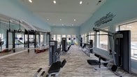 This dedicated fitness center is designed to support a comprehensive workout experience in a bright, open environment. The space features soaring ceilings and large windows along the walls, flooding the room with natural light and offering views of the surrounding outdoor landscape. The light blue walls create a calm, energizing atmosphere, complemented by motivational fitness-themed typography art displayed prominently on one wall — a fitting touch that reinforces the space's purpose.

The layout is thoughtfully organized to accommodate a variety of exercise styles. A diverse selection of strength training machines is arranged throughout the room, including cable machines, weight benches, and multi-use resistance equipment. Stability balls and additional accessories are stored neatly along the perimeter, supporting flexibility and functional training. The wood-look flooring adds a clean, modern aesthetic while providing a durable surface suited to high-traffic use.

Recessed lighting throughout the ceiling supplements the natural light, ensuring the space is well-lit at any time of day. The open floor plan allows for ease of movement between equipment, making the fitness center practical and accessible. Overall, this space delivers a well-rounded facility ideal for strength training, cardio, and general fitness routines.