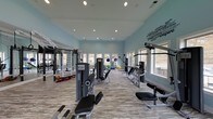 This dedicated fitness center is designed to support a comprehensive workout experience in a bright, open environment. The space features soaring ceilings and large windows along the walls, flooding the room with natural light and offering views of the surrounding outdoor landscape. The light blue walls create a calm, energizing atmosphere, complemented by motivational fitness-themed typography art displayed prominently on one wall — a fitting touch that reinforces the space's purpose.

The layout is thoughtfully organized to accommodate a variety of exercise styles. A diverse selection of strength training machines is arranged throughout the room, including cable machines, weight benches, and multi-use resistance equipment. Stability balls and additional accessories are stored neatly along the perimeter, supporting flexibility and functional training. The wood-look flooring adds a clean, modern aesthetic while providing a durable surface suited to high-traffic use.

Recessed lighting throughout the ceiling supplements the natural light, ensuring the space is well-lit at any time of day. The open floor plan allows for ease of movement between equipment, making the fitness center practical and accessible. Overall, this space delivers a well-rounded facility ideal for strength training, cardio, and general fitness routines.