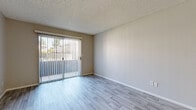 This single-floor residence offers a clean, functional layout with a cohesive design carried throughout. Light wood-style laminate flooring runs across the main living areas, complemented by neutral-toned walls and white baseboards that create a bright, airy atmosphere. The living room is an open, well-proportioned space anchored by a sliding glass door with vertical blinds that provides direct access to an outdoor patio and fills the room with natural light. A pendant-lit dining area flows seamlessly toward the kitchen, which is designed in a classic galley style. The kitchen features white cabinetry on both sides, dark countertops, white appliances including a range and refrigerator, and a sink positioned beneath a window. A pass-through counter connects the kitchen to the dining and living areas. The bedroom spaces are generously sized, with one room featuring an exposed concrete block wall that adds an industrial character, along with a closet equipped with a rod for hanging storage. Additional bedrooms offer windows with outdoor views and ample floor space. A hallway leads to the bathroom, which includes white vanity cabinetry, dark countertops, a large mirror, and a row of vanity lighting. Textured ceilings are present throughout, adding a traditional finish to the space.