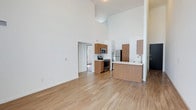 This two-story, 2-bedroom, 1-bathroom residence offers a thoughtfully designed open-concept layout with a striking double-height ceiling in the main living area, creating an expansive, airy atmosphere. The living space is bathed in natural light and features warm-toned hardwood-style flooring that flows seamlessly throughout. The kitchen is equipped with stainless steel appliances — including a range, microwave, and refrigerator — and features medium-toned wood cabinetry paired with a clean white countertop peninsula that doubles as a breakfast bar and divides the kitchen from the living area. The minimalist white walls complement the modern aesthetic, allowing the natural materials and stainless finishes to stand out. A hallway visible from the main living space leads to additional rooms, suggesting a functional and well-organized floor plan across both levels. The two bedrooms provide comfortable private retreats, while the bathroom is conveniently accessible. The overall ambiance of the space is clean, contemporary, and welcoming, making it well-suited for those who appreciate modern design, open living spaces, and practical, efficient layouts.