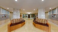 This building lobby presents a refined and welcoming environment defined by its polished terrazzo flooring, which features a striking starburst mosaic inlay as a central focal point. Tall marble-clad columns flank the main corridor, lending the space a sense of grandeur and structural elegance. Recessed lighting throughout ensures consistent, well-distributed illumination across the entire lobby.

Two generously sized curved benches with patterned upholstery and warm wood bases line the central corridor, providing comfortable seating for residents and visitors. A dedicated lounge alcove off the main hall is furnished with a pair of leather club chairs, a decorative wall clock, artwork, and a vase with floral accents, creating an intimate and welcoming sitting area.

The mailroom area features an extensive bank of individual mailboxes set against travertine-style wall panels, accompanied by an additional curved bench for convenience. Frosted glass-paneled windows above the mailboxes allow soft, diffused light into the area.

An elevator vestibule houses two side-by-side elevators with brushed metallic doors, offering smooth access throughout the building. The main entrance features glass doors that provide views to the surrounding greenery outside. Corridors extend from the central lobby, finished with consistent flooring and artwork, maintaining a cohesive and polished aesthetic throughout.