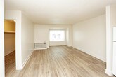 This single-floor residential unit offers a practical and well-organized layout designed for comfortable everyday living. The kitchen is equipped with warm medium-toned wood cabinetry spanning both upper and lower sections, a double-basin stainless steel sink, a white electric range, and a full-size refrigerator. Light-toned laminate wood flooring runs throughout, providing a cohesive and easy-to-maintain surface across the space. A textured popcorn ceiling is present in the kitchen and living areas, adding a classic touch to the interior. The living area is open and spacious, benefiting from a large window that invites natural light and creates an airy atmosphere. A wall-mounted PTAC heating and cooling unit ensures year-round climate control. The room connects smoothly to the rest of the unit, with a hallway leading to additional spaces. The bathroom features a full-size tub and shower combination with a curved curtain rod, clean white surround, brushed nickel hardware and towel bars, a toilet, and a dark-finished vanity. Neutral wall tones throughout the unit contribute to a calm, move-in-ready ambiance that is both functional and inviting.