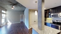 This single-floor space showcases a compelling blend of industrial and contemporary design elements that come together to create a stylish and functional living environment. The exposed concrete ceiling serves as a striking architectural focal point, lending an urban, loft-like character to the interior, while warm hardwood flooring throughout grounds the space with richness and continuity.

The open-concept layout seamlessly connects the kitchen and living areas, promoting a sense of spaciousness and ease of movement. The kitchen is appointed with granite countertops that offer both visual appeal and practicality, complemented by dark cabinetry with glass-front upper panels and vibrant blue lower cabinets that add a bold design accent. A pendant light fixture hangs above the counter, providing focused task lighting and enhancing the modern aesthetic.

A structural concrete column adds to the industrial character of the space while defining the transition between the kitchen and the living area. The living space features neutral gray walls and a ceiling fan, creating a comfortable and versatile area. A hallway leads toward additional rooms, with a set of paneled double doors providing access to a closet or utility space. Natural light filters throughout, contributing to a bright and welcoming ambiance across the entire floor.