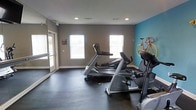 This fitness center is a dedicated single-floor space designed with functionality and motivation in mind. The room features a clean, open layout with dark rubberized flooring that provides both durability and traction during workouts. Natural light streams in through multiple windows along the wall, creating a bright and energizing atmosphere. The neutral gray walls are complemented by a bold teal accent wall, adding a dynamic visual element to the space. A decorative bicycle mounted on the accent wall serves as a creative focal point. The fitness area is equipped with quality cardio machines, including Matrix-brand treadmills and an elliptical trainer, offering residents a variety of aerobic exercise options. A floor-to-ceiling mirror along one wall enhances the sense of space and allows users to monitor their form during exercise. Recessed lighting in the ceiling supplements the natural light, ensuring the room remains well-lit at all times. Access is provided through a glass-paneled door, contributing to an open and welcoming feel. Overall, the space strikes a balance between practicality and aesthetic appeal, making it a functional and inviting environment for fitness enthusiasts.