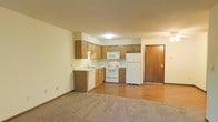 This single-floor space offers a practical and functional open layout, with the kitchen and living area seamlessly connected. The kitchen is equipped with medium-toned wooden cabinetry that provides generous storage along both upper and lower sections. White appliances, including a refrigerator, electric range, microwave, and dishwasher, are neatly integrated into the layout. A double-basin sink is positioned along the counter, and light-toned countertops complement the warm wood cabinetry throughout. The kitchen transitions naturally into the living area, which features durable hardwood-style flooring near the kitchen and soft carpeting in the main living zone, creating a comfortable and welcoming atmosphere. Neutral wall tones throughout contribute to a clean, understated aesthetic. A solid wood entry door is visible near the kitchen, providing a straightforward and accessible entry point into the unit. Ceiling-mounted lighting fixtures illuminate the kitchen area, ensuring adequate brightness. Overall, the space presents a straightforward, move-in-ready layout well-suited for everyday living.