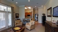 This single-floor residence offers a thoughtfully designed layout that seamlessly connects the living and kitchen areas. The living room is comfortably furnished with plush upholstered armchairs and a leather ottoman, creating a welcoming gathering space. Recessed lighting and crown molding add a refined architectural touch to the interior, lending an air of classic elegance throughout.

The kitchen is visible from the living area and features warm, honey-toned wood cabinetry topped with decorative accents. A raised bar-height counter with seating for two provides a casual dining option, making the space ideal for both everyday use and entertaining. Black appliances, including a built-in microwave, complement the cabinetry's warm tones.

The open sightlines between rooms enhance the sense of spaciousness, while neutral wall tones and carpeted flooring in the living area create a cozy, inviting ambiance. A corridor leading toward additional rooms hints at a well-organized floor plan beyond the main living space. Decorative wall art and carefully placed greenery add personal character to the home. Overall, this residence balances functional design with comfortable, understated style.