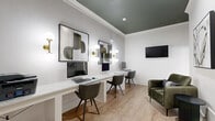 Located at 4045 George Busbee Pkwy NW in Kennesaw, Georgia, this Cyber Café office is a thoughtfully designed shared workspace that blends functionality with contemporary style. The single-room layout features light wood-look flooring and crisp white walls accented by elegant crown molding and a striking deep green ceiling that adds a sophisticated touch of color contrast.

A long, built-in white desk runs along one wall, accommodating multiple workstations equipped with desktop computers and ample surface space for productivity. Stylish dark olive-green upholstered chairs complement the room's refined color palette. Globe-shaped sconces with brushed brass fixtures are mounted along the desk wall, providing warm, focused task lighting while also serving as decorative elements. A wall-mounted flat-screen television is positioned on an adjacent wall for added functionality.

Curated abstract artwork in black frames adds a gallery-like quality to the space. A comfortable green velvet lounge chair and a sleek side table offer a relaxed seating area for breaks or informal meetings. A glass-paned interior door connects the office to an adjoining space, allowing natural light to filter through. The overall ambiance is polished, modern, and inviting — well-suited for focused work or casual connectivity.