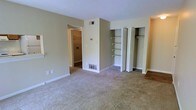 This single-floor space is finished in neutral beige tones throughout, creating a calm and cohesive atmosphere. The main living area features wall-to-wall carpeting that adds warmth underfoot, complemented by clean white baseboards and a textured ceiling. HVAC vents are conveniently positioned both on the wall and floor, ensuring climate control throughout the space.

A standout feature of this room is the built-in open shelving unit, offering generous storage and display space without occupying additional floor area. Adjacent to the shelving, a closet with bifold doors provides enclosed storage, keeping the space organized and tidy. Together, these storage solutions make the room highly functional.

A doorway leads to an adjoining room, visible in the background, which also features carpeting and appears to include additional closet or utility storage, suggesting flexible use of the connected spaces.

The layout is straightforward and practical, with ample open floor space that allows for versatile furniture arrangements. The neutral color palette and clean finishes throughout give the space a move-in-ready quality, well-suited for a variety of living arrangements.