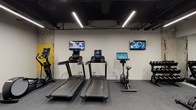 This fitness center offers a comprehensive workout environment thoughtfully designed to accommodate a wide range of exercise needs. The space features an industrial aesthetic, characterized by an exposed black ceiling with visible ductwork and structural columns that retain their original raw, textured finish with distinctive yellow accents — lending the gym an urban, loft-like character.

Underfoot, durable rubber flooring provides safe and comfortable traction throughout the space. Linear LED strip lighting runs across the ceiling, delivering bright, even illumination across the entire floor.

The cardio zone is well-stocked with multiple treadmills and stationary bikes, each equipped with individual digital display screens. Wall-mounted flat-screen televisions are positioned throughout this area, allowing users to stay entertained during their workouts.

The strength training area features multiple dumbbell racks with a full range of free weights, adjustable workout benches, and a multi-functional cable machine. A large floor-to-ceiling mirror spans one wall, enhancing the sense of space while allowing users to monitor their form.

Additional equipment includes an elliptical trainer and a stair climber, rounding out a diverse selection of fitness options. A medicine ball rack and battle ropes add functional training capabilities. The space also includes a dedicated utility room accessible via a clearly marked door.