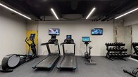 This fitness center offers a comprehensive workout environment thoughtfully designed to accommodate a wide range of exercise needs. The space features an industrial aesthetic, characterized by an exposed black ceiling with visible ductwork and structural columns that retain their original raw, textured finish with distinctive yellow accents — lending the gym an urban, loft-like character.

Underfoot, durable rubber flooring provides safe and comfortable traction throughout the space. Linear LED strip lighting runs across the ceiling, delivering bright, even illumination across the entire floor.

The cardio zone is well-stocked with multiple treadmills and stationary bikes, each equipped with individual digital display screens. Wall-mounted flat-screen televisions are positioned throughout this area, allowing users to stay entertained during their workouts.

The strength training area features multiple dumbbell racks with a full range of free weights, adjustable workout benches, and a multi-functional cable machine. A large floor-to-ceiling mirror spans one wall, enhancing the sense of space while allowing users to monitor their form.

Additional equipment includes an elliptical trainer and a stair climber, rounding out a diverse selection of fitness options. A medicine ball rack and battle ropes add functional training capabilities. The space also includes a dedicated utility room accessible via a clearly marked door.