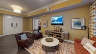 This thoughtfully designed single-floor community space offers a variety of interconnected areas tailored for relaxation, dining, and social interaction. The main lounge area features a warm, inviting atmosphere with a mix of plush armchairs upholstered in rich earth tones—rust orange, striped multi-color, and deep brown leather—arranged around low coffee tables atop patterned carpeting. A stone accent wall with a wall-mounted flat-screen TV serves as a focal point, anchored by elegant white columns and overhead drum pendant lighting that bathes the space in a soft, ambient glow. Curated artwork adorns the neutral gray walls throughout, adding visual interest and a sense of refinement. A cozy sitting nook offers additional seating with a round side table and floor lamp, ideal for quiet conversation or reading. The fully equipped kitchen is outfitted with dark cabinetry, stainless steel appliances, a tile backsplash, and a generous island with granite countertops and bar-height seating beneath pendant lights—perfectly suited for communal dining and cooking activities. Adjacent to the kitchen, the dining room provides ample seating at multiple tables with coordinating upholstered chairs and wall-mounted televisions, creating a welcoming environment for shared meals and group gatherings. Dark hardwood flooring flows throughout the dining and kitchen areas, unifying the space with a polished, contemporary finish.