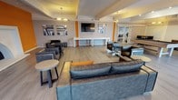 This single-floor space is designed as a contemporary lounge and recreation area, offering a well-appointed environment for socializing and entertainment. The main lounge area features generous seating arrangements, including large leather-style sofas and armchairs in a dark gray tone, complemented by orange accent pillows that echo the bold orange vertical wall accents throughout the space. Light wood-look flooring runs throughout, contributing to a clean, modern aesthetic.

A wall-mounted flat-screen television serves as a focal point on the main wall, flanked by abstract geometric artwork that reinforces the space's contemporary design theme. Beneath the TV, a white shuffleboard table provides a central recreational feature. Pendant lighting and overhead fixtures offer warm, ambient illumination across the area.

The layout flows openly, with clearly defined seating clusters that encourage conversation. A secondary area is visible beyond the main lounge, suggesting additional seating or activity zones separated by glass-paneled partitions. The overall ambiance is modern and inviting, with a cohesive color palette of gray, white, and orange creating a vibrant yet comfortable communal environment.