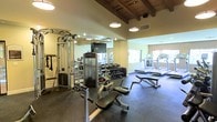 This fitness center combines functional design with an inviting atmosphere, making it a well-rounded space for a variety of workout routines. The ceiling is a standout architectural feature, showcasing exposed wooden beams that lend a warm, rustic character to the room, complemented by modern drum pendant lights that provide even, comfortable illumination throughout the space.

The flooring is covered in durable rubber matting, ideal for both heavy equipment and high-impact exercise. Large mirrors line the walls, visually expanding the space and allowing users to monitor their form during workouts. A flat-screen television is mounted for entertainment during exercise sessions.

The strength training area is well-appointed, featuring a multi-station cable machine, a dedicated free weight rack stocked with a comprehensive range of dumbbells, and an adjustable workout bench. The cardio section is equipped with multiple treadmills positioned near windows, which allow natural light to enter and provide an open, airy feel. The overall ambiance is clean, organized, and functional, creating an environment well-suited for both casual and dedicated fitness enthusiasts.