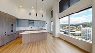 Located at 1510 SW Alder in Portland, Oregon, this contemporary residence delivers an impressive blend of modern design and urban living. Soaring double-height ceilings define the open-concept living and kitchen area, amplifying the sense of space and allowing natural light to pour in through expansive floor-to-ceiling windows. The living area opens directly onto a private balcony through wide sliding glass doors, offering sweeping panoramic views of the Portland cityscape and surrounding tree-lined hills.

The kitchen is thoughtfully appointed with matte blue-gray upper cabinetry, warm wood-toned lower cabinets, a striking marble-style backsplash, white quartz countertops, a large center island with a waterfall edge, and sleek black hardware throughout. Pendant lighting and a full-size panel-front refrigerator complete the refined aesthetic. Light hardwood flooring flows throughout the main living spaces.

The bedroom is generously sized with soft carpet, tall ceilings, and access to a well-organized walk-in closet featuring built-in shelving and a drawer unit. The primary bathroom offers a dual-sink vanity with quartz counters, wood-toned cabinetry, matte black fixtures, and a marble-tiled walk-in shower. A secondary bathroom mirrors the same refined finishes with a glass-enclosed shower and clean-lined vanity.