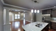 This single-floor residence showcases an open-concept layout that seamlessly connects the kitchen and living areas, creating a sense of spaciousness and flow. The kitchen is anchored by a large granite-topped island with a double stainless steel sink and modern pendant lighting — three sleek white fixtures suspended from the ceiling — that add both function and style. Dark cabinetry and built-in appliances complement the light-toned granite countertops, striking a sophisticated contrast.

The transition into the living area is defined by elegant decorative columns and rich hardwood flooring that extends throughout the space. Crown molding runs consistently along the ceilings, adding a refined architectural detail. The living room features a distinctive tray ceiling with a ceiling fan at its center, lending depth and character to the room. Neutral warm-gray walls provide a versatile backdrop that works well with a variety of interior styles.

Natural light filters in through multiple windows, brightening the living area and enhancing the warmth of the hardwood floors. The overall ambiance of the space is polished yet inviting, with thoughtful architectural details — from the column accents to the layered ceiling work — that elevate the design throughout.