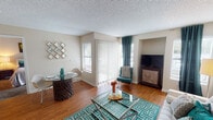 This single-floor residence offers a thoughtfully designed layout that balances comfort and functionality throughout. The living area serves as the heart of the home, showcasing warm hardwood flooring, a textured ceiling, and an abundance of natural light streaming in through large windows and a sliding glass door dressed with vertical blinds and teal accent curtains. A built-in nook provides a dedicated media area, while the open layout seamlessly accommodates both a sitting area and a dining space. The kitchen features crisp white cabinetry, light stone-finish countertops, a stainless steel sink, and under-cabinet lighting, creating a clean and practical workspace. The bedroom is carpeted for comfort and benefits from direct outdoor access via a sliding glass door with vertical blinds, allowing ample natural light. A dedicated hallway off the bedroom leads to a well-lit bathroom with white vanity cabinetry, a mirrored medicine cabinet, and warm overhead lighting, alongside a separate walk-in closet with built-in shelving. Throughout the residence, neutral warm-toned walls, consistent trim work, and cohesive teal and neutral décor accents contribute to a welcoming and harmonious ambiance.