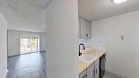 This single-floor unit has been thoughtfully renovated with a clean, modern aesthetic carried consistently throughout every room. The kitchen serves as a functional centerpiece, featuring gray shaker-style cabinetry with matte black hardware, warm butcher block countertops, a classic white subway tile backsplash, a deep white farmhouse sink with a sleek matte black pull-down faucet, and a dishwasher. A pass-through counter opening connects the kitchen to an adjacent dining or living area, enhancing the sense of openness between spaces. The living area is bright and airy, benefiting from a large sliding glass door that opens to a private balcony, allowing natural light to fill the room. Consistent wood-look plank flooring in a cool gray tone flows throughout the entire unit, providing a cohesive and contemporary feel underfoot. The bedroom offers a comfortable, well-proportioned layout with a window fitted with blinds for privacy and light control. The bathroom features matching gray cabinetry with a white countertop and sink, matte black hardware, tile flooring, and an industrial-style pendant light. Textured ceilings are present throughout, and crisp white walls provide a neutral canvas across all rooms.