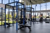 Located at 2700 W Cobalt Dr in Meridian, Idaho, this two-story fitness facility is designed to deliver a comprehensive workout experience within a modern, light-filled environment. The architectural highlight is the ground-floor weight room, which rises to a dramatic double-height ceiling framed by floor-to-ceiling windows. This soaring space floods the room with natural light and offers open views of the surrounding grounds and community, creating an energizing atmosphere. The weight room is equipped with power racks, Rogue-branded strength equipment, adjustable benches, and a full array of free weights, providing a serious training environment for strength athletes and general fitness users alike. A dedicated cardio area features treadmills, ellipticals, a rowing machine, and cable machines, all positioned along windowed walls to take advantage of natural light and outdoor scenery. A separate studio space is distinguished by teal accent walls, a wall-mounted display screen, and open floor space suitable for group fitness, cycling, or functional training. The second floor overlooks the main weight room through large glass partitions and metal-railed mezzanine walkways, maintaining visual connectivity between levels while housing additional cardio equipment with panoramic views of the outdoor pool area and surrounding landscape. Throughout both floors, pendant lighting, coffered ceilings, and neutral flooring contribute to a clean, contemporary aesthetic.