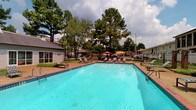 This amenity area features a large outdoor swimming pool with clear turquoise water, set within a spacious pool deck finished in brick-toned pavers. The deck is furnished with multiple lounge chairs and sun loungers positioned along the pool's perimeter, along with colorful patio umbrellas providing shaded seating areas for relaxation. Tall, mature pine trees frame the space, offering a natural backdrop and partial shade throughout the day. A fenced perimeter ensures privacy and security for the area.

Adjacent to the pool is a dedicated fitness center, accessible through a glass-paneled entry door clearly labeled "Fitness Center." The gym features large windows that allow natural light to flood the interior and offer direct views of the pool area. Inside, the space is equipped with a row of treadmills, weight training equipment, free weights, and exercise balls, all set on patterned carpet flooring. A small tiled entryway connects the fitness center to the outdoor amenity space, providing a clean transition between the indoor and outdoor environments. Together, these amenities form a well-rounded recreational area designed for both activity and leisure.