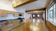 This single-floor loft showcases an open-concept layout that seamlessly blends industrial character with modern functionality. The space is defined by its striking exposed brick walls, which add warmth and texture throughout. Overhead, heavy timber beams span the ceiling, lending a rustic, warehouse-inspired aesthetic that complements the polished hardwood floors below, which reflect light and enhance the sense of openness.

The kitchen is positioned along one wall and features light maple cabinetry, dark granite countertops, and stainless steel appliances including a gas range, built-in microwave, and a sink set into an island-style counter that opens toward the main living area. Track lighting runs along the ceiling beams, providing adjustable illumination across the expansive open floor plan.

The living area extends generously from the kitchen, framed by additional exposed brick and original timber columns that serve as architectural focal points. Windows set within the brick walls allow natural light to filter in, brightening the interior while preserving the raw, loft-style character. The overall ambiance is one of spacious, industrial elegance with practical, contemporary finishes.