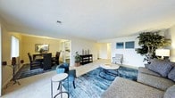 This single-floor space is designed around an open-concept layout that promotes a seamless connection between the living, dining, and kitchen areas. The living room serves as the central gathering space, featuring wall-to-wall carpeting in a neutral beige tone and a textured ceiling that adds subtle dimension overhead. A bold blue area rug anchors the seating arrangement, which includes a round coffee table and accent chairs, creating a defined yet open lounge area. Along one wall, a dark console table provides functional storage and display space, accompanied by potted plants that introduce a natural element to the interior.

A wide passthrough opening leads into the dining area, which is illuminated by pendant lighting overhead, lending warmth to the space. The kitchen is visible beyond, finished in light tones with white cabinetry and standard appliances. To the right of the living room, an arched hallway opening suggests access to additional rooms within the unit. The overall palette of warm neutrals, accented by blues and greens, gives the space a calm, livable atmosphere suited for both everyday comfort and casual entertaining.