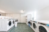 Located at 30 Gilder Dr in Toronto, Ontario, this shared laundry facility offers a clean, well-organized space designed to accommodate the laundry needs of multiple residents efficiently. The room is finished with white tile-clad walls and durable grey-green ceramic floor tiles throughout, creating a bright and hygienic environment that is easy to maintain.

The facility is generously equipped with a row of front-loading washing machines along one wall, paired with both stacked and standalone dryers to maximize capacity. Each washer features individual water supply connections with shutoff valves, and a payment kiosk is conveniently positioned in the center of the room for easy access. Ample folding space is provided via long countertops with dark work surfaces, including a dedicated utility sink area ideal for pre-treating garments or hand-washing delicate items.

Overhead fluorescent and ceiling-mounted light fixtures ensure the space is well-lit throughout. A ventilation unit is installed along the upper wall to maintain proper airflow. A bulletin board with posted notices keeps residents informed of facility rules and updates. The layout is thoughtfully arranged to allow comfortable movement between machines and folding areas, making this laundry room a practical and functional shared amenity.