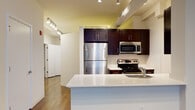 This single-floor residence offers a thoughtfully designed open-concept layout that seamlessly connects the kitchen and living areas. The kitchen is a standout feature, anchored by a sleek white quartz island with an integrated sink, providing ample prep space and a natural gathering point. Rich espresso-toned cabinetry extends to the upper walls, offering generous storage, while a full suite of stainless steel appliances — including a full-size refrigerator, range with electric cooktop, and built-in microwave — delivers a polished, modern aesthetic. Track lighting runs throughout the kitchen and hallway, ensuring consistent, well-distributed illumination across the space. The living area is bathed in natural light through a pair of large, black-framed windows that frame views of the surrounding urban environment. Warm hardwood flooring flows continuously throughout the main living spaces, adding depth and cohesion to the interior. A hallway connects the main living zone to additional rooms, including a bathroom appointed with a dark-cabinet vanity, white countertop, brushed nickel fixtures, and a large mirror. The overall design is clean and contemporary, with white walls and structural beam details that lend an urban character to the residence.