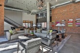 Located at 915 Glenwood Ave SE in Atlanta's vibrant Glenwood Park neighborhood, this single-floor community lounge delivers an immersive industrial-chic experience. The space is defined by soaring double-height ceilings with exposed ductwork and structural brick walls that lend a raw, warehouse-inspired character throughout. Floor-to-ceiling steel-framed windows flood the interior with abundant natural light while offering open views of the surrounding outdoor area and pool terrace.

One of the most distinctive architectural features is the series of large concrete tunnel-pod seating alcoves set against the exposed brick wall. Each pod frames a cozy nook with built-in booth seating and a private TV, creating intimate gathering spaces within the expansive open floor plan. Complementing these pods, the main lounge area is furnished with richly upholstered marble-patterned chairs, dark leather sofas, and warm accent lamps beneath a dramatic sculptural chandelier.

A rustic wood-and-concrete wet bar island anchors the social hub of the space, complete with bar seating and beverage storage. A decorative display of mounted skateboards adorns one brick wall, adding a local, urban artistic flair. An open staircase with metal railings connects to an upper mezzanine level. Polished concrete floors tie the entire space together, reinforcing the modern industrial aesthetic throughout.