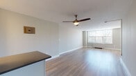 Located at 425 Dayton Towers Dr in Dayton, Ohio, this single-floor unit offers a clean, modern layout designed for comfortable everyday living. The open living and dining area is a standout feature, showcasing light-toned hardwood flooring that runs throughout the space, neutral gray walls, and a ceiling fan with a brass accent light fixture. A large window at the far end of the living area fills the room with natural light and provides views of the surrounding tree line, contributing to a bright and airy atmosphere. A baseboard heating unit is positioned beneath the window for year-round comfort. The galley-style kitchen is efficiently designed with white cabinetry, gray quartz countertops, a stainless steel sink, subway tile backsplash, and black appliances including a gas range, refrigerator, and dishwasher. The bathroom is tastefully updated with large-format gray tile covering the walls, a white vanity with granite-style countertop, brushed nickel fixtures, a full tub and shower combination, and a wide vanity mirror with overhead lighting. Additional rooms provide flexible living space, each reflecting the same clean, neutral aesthetic with consistent hardwood flooring and white trim throughout.