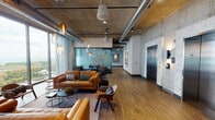 This striking amenity floor showcases a seamless blend of industrial and contemporary design, defined by exposed concrete walls and ceilings, visible ductwork, and warm wood-plank flooring throughout. Soaring double-height ceilings anchor a dramatic main lounge adorned with a vibrant gallery wall of large-scale colorful artwork, complemented by an eclectic mix of leather and upholstered seating arranged around sleek black coffee tables. Cascading glass pendant lights in varying shapes and amber hues add warmth and visual interest across the open-plan layout.

Floor-to-ceiling windows flood the space with abundant natural light while framing sweeping views of the surrounding urban skyline. A glass-enclosed conference room offers a private meeting setting with raw concrete walls, curated artwork, and direct access to city views. The social lounge and game area features a pool table, casual dining tables, a built-in bar counter with dual ovens, mounted flat-screen TVs, and a bold geometric-tiled accent wall in yellow and white.

A serene outdoor terrace, accessible through full-height sliding glass doors, provides an open-air retreat with modern seating and planted trees set against the cityscape. Frosted glass partitions and sliding barn-style doors define additional flex spaces, while elevator access and smart technology panels enhance the overall functionality of this well-appointed, design-forward amenity floor.
