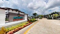 Monarch Station is a contemporary residential community that makes a striking first impression with its well-appointed gated entrance at 4901. The community entrance features a bold, eye-catching monument sign with vibrant teal and multicolored branding set against a clean, modern stucco and tile facade. The entry approach is paved with attractive decorative brick pavers, complemented by meticulously maintained tropical landscaping that includes tall palm trees, colorful bromeliads, and lush ground cover plantings in vivid shades of red and green. A controlled-access gate provides security at the entryway, leading into the broader community beyond. The multi-story residential buildings visible in the background reflect a modern architectural style, characterized by clean lines, neutral color palettes, and a well-organized layout. The overall ambiance of the community entrance conveys a sense of polished, resort-style living with a strong tropical Florida aesthetic. The combination of thoughtful landscaping, decorative hardscaping, and contemporary architecture establishes Monarch Station as a well-designed and visually appealing residential destination.