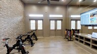 This single-story fitness room combines functional design with a clean, modern aesthetic. The interior walls are finished in horizontal shiplap paneling painted in a warm neutral tone, complemented by an exposed brick accent wall that adds texture and visual interest. Light wood-look flooring runs throughout, contributing to a bright, welcoming atmosphere.

The room is equipped with cycling bikes positioned along one side, alongside a well-stocked dumbbell rack displaying a full range of colorful free weights, making the space suitable for both cardio and strength training. A ceiling fan is centrally mounted overhead, providing consistent air circulation during workouts.

Natural light enters generously through multiple glass-paneled doors and clerestory windows positioned high along the walls, illuminating the space without compromising wall area needed for equipment. The elevated windows also enhance the sense of height within the room.

The double-door entry opens outward, offering convenient access and ventilation. Overall, the space balances practicality with thoughtful design details, creating an inviting environment well-suited for group or individual fitness activities.