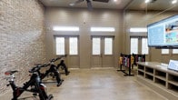 This single-story fitness room combines functional design with a clean, modern aesthetic. The interior walls are finished in horizontal shiplap paneling painted in a warm neutral tone, complemented by an exposed brick accent wall that adds texture and visual interest. Light wood-look flooring runs throughout, contributing to a bright, welcoming atmosphere.

The room is equipped with cycling bikes positioned along one side, alongside a well-stocked dumbbell rack displaying a full range of colorful free weights, making the space suitable for both cardio and strength training. A ceiling fan is centrally mounted overhead, providing consistent air circulation during workouts.

Natural light enters generously through multiple glass-paneled doors and clerestory windows positioned high along the walls, illuminating the space without compromising wall area needed for equipment. The elevated windows also enhance the sense of height within the room.

The double-door entry opens outward, offering convenient access and ventilation. Overall, the space balances practicality with thoughtful design details, creating an inviting environment well-suited for group or individual fitness activities.