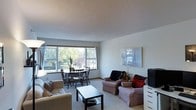 This single-floor space presents an open-concept layout where the living and dining areas flow seamlessly into one another. The living room is furnished with cushioned armchairs and a sleek black coffee table, centered on neutral carpeted flooring that extends throughout the space. A textured popcorn ceiling adds a classic touch overhead, while the walls are kept in a clean, neutral white tone that enhances the sense of openness.

A standout feature of the space is the large, multi-pane window that spans much of one wall in the dining area, drawing in generous natural light and offering pleasant views of mature, leafy trees outside. This window creates a bright and airy atmosphere, making the combined living and dining area feel welcoming and expansive. The dining area comfortably accommodates a round wooden table with matching chairs, positioned to take full advantage of the outdoor view.

A framed piece of wall art accents the living area, while a compact storage unit provides practical organization near the seating arrangement. Overall, the space offers a functional and comfortable single-level layout with a light-filled, relaxed ambiance.