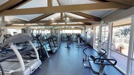 This fitness center is housed in a single-floor structure distinguished by its striking vaulted ceiling with exposed wooden beam trusses, lending the space an open, airy character. The high-pitched roofline creates a generous sense of vertical space, while recessed and track lighting ensures the interior is well-illuminated at all hours. Large windows line the perimeter walls, inviting abundant natural light and offering views of the surrounding greenery, contributing to an energizing workout atmosphere. The flooring throughout is a durable dark rubber mat, well-suited for high-traffic exercise use. The cardio section is comprehensively outfitted with elliptical trainers, treadmills, and stationary bikes, providing a full range of aerobic options. Strength training equipment is arranged along the mirrored wall at the far end of the room, with the mirrors effectively amplifying the sense of space and allowing users to monitor their form. The layout is thoughtfully organized to allow comfortable movement between stations. A glass-paned door provides convenient access to the outdoors. Overall, the space strikes a balance between functional fitness facility design and an inviting, naturally lit environment.