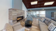 This single-floor communal space is thoughtfully designed to offer a comfortable and functional environment across several distinct areas. The kitchen is a standout feature, equipped with stainless steel appliances including a refrigerator, electric range, and built-in microwave. Rich mahogany cabinetry pairs elegantly with granite countertops, while a breakfast bar with a white brick base and bar seating provides a casual dining option. Light hardwood flooring runs throughout, tying the space together cohesively. The lounge area is anchored by a linear gas fireplace set into a wood-panel accent wall, creating a warm and inviting atmosphere. A striking coffered ceiling with dark wood panels adds architectural depth and visual interest overhead. Comfortable seating arrangements make this area well-suited for relaxed gatherings. A dedicated TV room offers a wall-mounted flat-screen television flanked by black leather seating and a wood coffee table, providing a more private retreat. The dining area is flooded with natural light through expansive floor-to-ceiling sliding glass doors and windows that frame views of the surrounding outdoor landscape. Multiple dining tables accommodate groups of various sizes. Together, these areas create a well-rounded, multi-functional space ideal for both socializing and everyday living.