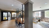 The Wellington Clubhouse in Arlington, Virginia offers a thoughtfully designed community gathering space that blends contemporary aesthetics with a warm, inviting atmosphere. The single-floor layout encompasses two distinct zones that flow seamlessly into one another, unified by light hardwood flooring, textured gray grasscloth-style wall coverings, and recessed lighting throughout.

The lounge area is anchored by a set of tufted leather sofas and mid-century accent chairs in rich cognac tones, arranged around a low coffee table atop a patterned area rug. Large windows dressed with sheer curtains allow natural light to filter in while offering views of the surrounding greenery. A wall-mounted flat-screen TV and curated artwork — including a bold black-and-white abstract piece and a framed DCA airport map print — add character to the space.

The dining and gathering zone features a substantial walnut communal table flanked by contemporary mixed chairs, illuminated by a striking row of black pendant lights suspended from the ceiling. Structural columns wrapped in matching textured material define the space without interrupting its open feel. A built-in kitchenette with white cabinetry, a beverage refrigerator, and geometric tile accents provides convenient amenities for residents and guests. A billiards table rounds out the entertainment offerings, making this clubhouse a well-appointed hub for community life.
