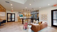 Located at 3529 Adirondack Way in Wilmington, NC, this single-floor community clubhouse is a thoughtfully designed gathering space that blends modern aesthetics with warm, eclectic décor. The open-plan lounge area serves as the heart of the space, featuring tufted leather seating, velvet ottomans, and a central bar-height table illuminated by a cluster of woven rattan pendant lights. A sleek black-framed glass partition wall separates zones while maintaining an open, airy feel throughout. Natural light pours in through tall windows dressed with dark curtain panels, complemented by a striking botanical-print accent wall and an ornate chandelier overhead. A curated gallery wall anchors one end of the lounge, incorporating framed artwork, a wall-mounted TV, and natural wicker accent chairs, adding a layered, personalized touch. The space transitions seamlessly into a private co-working area, where artisan rattan arch panels back individual workstations equipped with desktop computers and ergonomic seating. The tropical wallpaper motif carries through this section, creating a cohesive and inspiring work environment. Recessed lighting throughout ensures consistent illumination, while the neutral flooring and white walls allow the bold furnishings and décor to take center stage, resulting in an inviting and functional communal retreat.