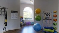 This single-floor fitness studio is thoughtfully designed to support a wide range of exercise and training activities. The space immediately impresses with its soaring ceilings and a striking arched window that floods the room with natural light and offers a pleasant view of surrounding greenery, creating an inviting and energizing atmosphere for workouts.

The interior features durable hardwood-style flooring, which provides both aesthetic appeal and a practical surface for a variety of exercises. The walls are finished in a clean, neutral tone, keeping the space feeling open and uncluttered. A workout reference poster is mounted on the wall, offering visual guidance for exercise routines.

The studio is well-stocked with functional training equipment, including a multi-tier kettlebell and medicine ball rack, stacked foam exercise step platforms, and a wall-mounted stability ball storage rack holding multiple large exercise balls in varying sizes. A resistance training station with cables and bands is also present, offering versatile upper and lower body workout options.

Overhead fluorescent lighting supplements the abundant natural light, ensuring the space remains well-lit at all times. The overall ambiance is clean, purposeful, and welcoming — well-suited for personal training sessions, group fitness classes, or individual exercise routines.