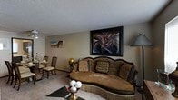 This multi-story residence spans three floors, offering a well-appointed layout designed for comfortable living. The living area serves as a focal point of the home, featuring warm, neutral-toned carpeting and textured ceilings that add depth and character to the space. A elegantly carved sofa with ornate wooden detailing and richly patterned cushions anchors the room, complemented by a dark glass-topped coffee table that adds a contemporary contrast to the traditional furnishings. Bold, large-scale wall art featuring vivid abstract imagery lends a dynamic visual element to the space, while smaller framed pieces contribute to a curated, gallery-like ambiance. A dining area is visible nearby, furnished with classic wooden chairs and a table, creating a seamless transition between living and dining spaces. The overall aesthetic blends traditional and eclectic design sensibilities, with warm earth tones, decorative accents including sculptural pieces and ornamental bowls, and layered textures throughout. The thoughtful arrangement of furniture and art creates an intimate yet visually rich environment, reflective of a home designed with both style and comfort in mind across all three of its floors.
