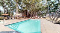 This outdoor amenity space centers around a large, rectangular swimming pool with clearly marked shallow and deep ends, offering a refreshing retreat surrounded by a generously sized concrete sundeck. Rows of sling-style lounge chairs line the poolside, providing ample space for relaxation under the open sky, while a shaded seating area with an umbrella-topped table offers a cooler alternative for outdoor gatherings. The entire pool area is enclosed by a sturdy dark metal security fence, ensuring safety and privacy for residents and guests.

Mature, towering trees frame the perimeter of the space, casting dappled shade and contributing to a serene, park-like atmosphere. A covered outdoor pavilion adjacent to the pool area showcases striking craftsman-style architecture, featuring exposed heavy timber beams, angled bracket supports, and a pitched gabled roof — all accented by natural wood tones and stone detailing. Carefully curated landscaping with flowering plants, ornamental grasses, and layered greenery lines the walkways and fencing, adding vibrant color and texture. The combination of thoughtful architectural design, lush natural surroundings, and well-furnished outdoor spaces creates an inviting and cohesive communal environment ideal for leisure and socializing.
