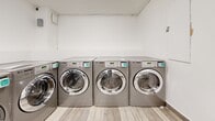 Located at 55 Bay St in Victoria, BC, this shared laundry facility is a compact yet efficiently organized single-floor space designed to serve a high volume of users. The room is laid out in an L-shaped configuration, making smart use of its available floor area.

A row of commercial-grade LG front-load washing machines lines the main wall, offering multiple units to accommodate simultaneous use. The machines feature digital displays and modern controls, suggesting a card or app-based payment system, as indicated by posted instructional signage on the wall. A separate bank of matching dryers is also present, ensuring a complete laundry experience.

The utility area includes a stainless steel work table, ideal for folding and sorting laundry, as well as a wall-mounted utility sink with an overhead faucet for pre-treatment or handwashing needs. An overhead wall-mounted air conditioning unit ensures year-round comfort for users.

The space is finished with light wood-look tile flooring throughout, contributing a clean and neutral aesthetic. Bright fluorescent strip lighting illuminates the entire area evenly. The walls are painted white, enhancing the sense of cleanliness and openness. Secure entry is provided via a solid grey door with a window panel, ensuring controlled access. Overall, the facility is practical, well-maintained, and efficiently designed for communal use.