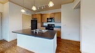 This single-floor residence offers a thoughtfully designed layout that balances comfort and functionality. The main living area features rich hardwood-style flooring, crown molding, and a ceiling fan with light fixture, creating a warm and inviting atmosphere. Multiple windows fitted with horizontal blinds allow natural light to fill the space, with views of the surrounding exterior. A door provides direct outdoor access from the living area.

The kitchen is a standout feature, centered around a large island with a double sink and dark countertops. Warm wood cabinetry lines the walls and provides ample storage, complemented by black appliances including a refrigerator, range, and microwave. Pendant and track lighting illuminate the workspace effectively. A built-in desk nook with open shelving and matching countertop adds a practical workspace just off the kitchen near the front entry.

The bedroom is finished with plush carpet and features a window with exterior views, offering a quiet and comfortable retreat. The hallway connecting the rooms is lined with hardwood-style flooring and includes a thermostat panel for climate control.

The full bathroom is well-equipped with a soaking tub framed by tile surround, a separate glass-enclosed shower with tile walls, a single-basin vanity with dark countertops, and tile flooring. A walk-in closet is accessible directly from the bathroom, adding generous storage to the space.
