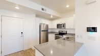 This thoughtfully laid-out one-bedroom, one-bathroom unit offers a clean and contemporary living experience across a single floor. The open-concept kitchen serves as a central feature, showcasing crisp white shaker-style cabinetry, grey quartz countertops, and a full suite of stainless steel appliances including a refrigerator, range, microwave, and dishwasher. A peninsula with bar-style overhang connects the kitchen to the rest of the living space, providing additional counter space and a natural division between cooking and lounging areas. Light wood-style flooring runs throughout the main living areas, lending a warm, modern aesthetic, while recessed lighting keeps the space bright and well-illuminated. The living room benefits from natural light through large windows fitted with horizontal blinds, and a glass door opens directly to a private balcony with outdoor views. The bedroom is carpeted for comfort and also features its own glass door access to the balcony, offering views of surrounding greenery. The bathroom is finished in neutral tones with tile flooring, a full tub-shower combination, and a vanity with a grey countertop. The overall ambiance is clean, modern, and functional, suited for comfortable everyday living.