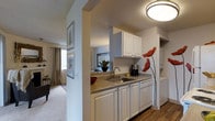 This single-floor residence is designed with an efficient, open layout that connects the kitchen directly to the living area, creating a cohesive and functional living environment. The kitchen features white raised-panel cabinetry on both upper and lower sections, providing ample storage space. A stainless steel sink sits beneath upper cabinets, accompanied by laminate countertops and a dishwasher integrated seamlessly into the cabinetry. A flush-mount ceiling light illuminates the kitchen area, while decorative wall art adds a touch of character to the space. The transition between the kitchen and living area is defined by a partial wall, which helps delineate the spaces while maintaining an open feel. The living area is finished with wall-to-wall carpeting and receives natural light through a window dressed with floor-length curtains. Framed artwork decorates the walls throughout, contributing to a cohesive aesthetic. The textured ceiling is consistent across both spaces, and the overall color palette of soft neutrals creates a clean, welcoming ambiance throughout the unit.