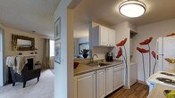 This single-floor residence is designed with an efficient, open layout that connects the kitchen directly to the living area, creating a cohesive and functional living environment. The kitchen features white raised-panel cabinetry on both upper and lower sections, providing ample storage space. A stainless steel sink sits beneath upper cabinets, accompanied by laminate countertops and a dishwasher integrated seamlessly into the cabinetry. A flush-mount ceiling light illuminates the kitchen area, while decorative wall art adds a touch of character to the space. The transition between the kitchen and living area is defined by a partial wall, which helps delineate the spaces while maintaining an open feel. The living area is finished with wall-to-wall carpeting and receives natural light through a window dressed with floor-length curtains. Framed artwork decorates the walls throughout, contributing to a cohesive aesthetic. The textured ceiling is consistent across both spaces, and the overall color palette of soft neutrals creates a clean, welcoming ambiance throughout the unit.