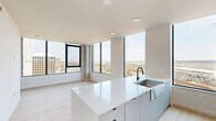 Located on the 13th floor at 3030 Potomac Ave in Arlington, VA, this contemporary 2-bedroom, 2-bathroom residence offers an open, light-filled layout with striking panoramic views of the surrounding Arlington skyline and beyond. Floor-to-ceiling windows with bold black frames define the living area, flooding the space with natural light and framing sweeping vistas that extend toward the horizon.

The open-concept kitchen and living area form the heart of the home, anchored by a large white quartz waterfall island with an undermount sink and matte black faucet. Light hardwood flooring runs throughout, complementing the crisp white walls and modern finishes.

The primary bedroom is generously proportioned, featuring expansive windows with unobstructed views and direct access to a private balcony through sleek sliding glass doors. The second bedroom also enjoys large windows with open city views, providing an equally bright and airy feel.

Both bathrooms are finished with modern fixtures and illuminated vanity mirrors. The primary en-suite stands out with vibrant blue large-format floor tiles, a glass-enclosed shower, and a window with an elevated view. The second bathroom features neutral tile flooring and clean white cabinetry. Each bedroom is accompanied by a dedicated closet, and a hallway connects the living spaces efficiently throughout this well-designed high-rise residence.